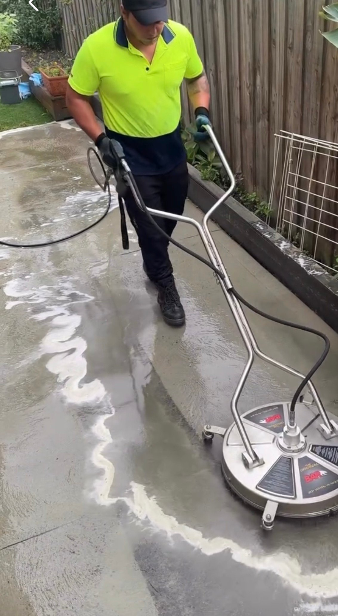 Worker in high-visibility shirt pressure cleaning a wet driveway with professional rotary surface cleaner equipment